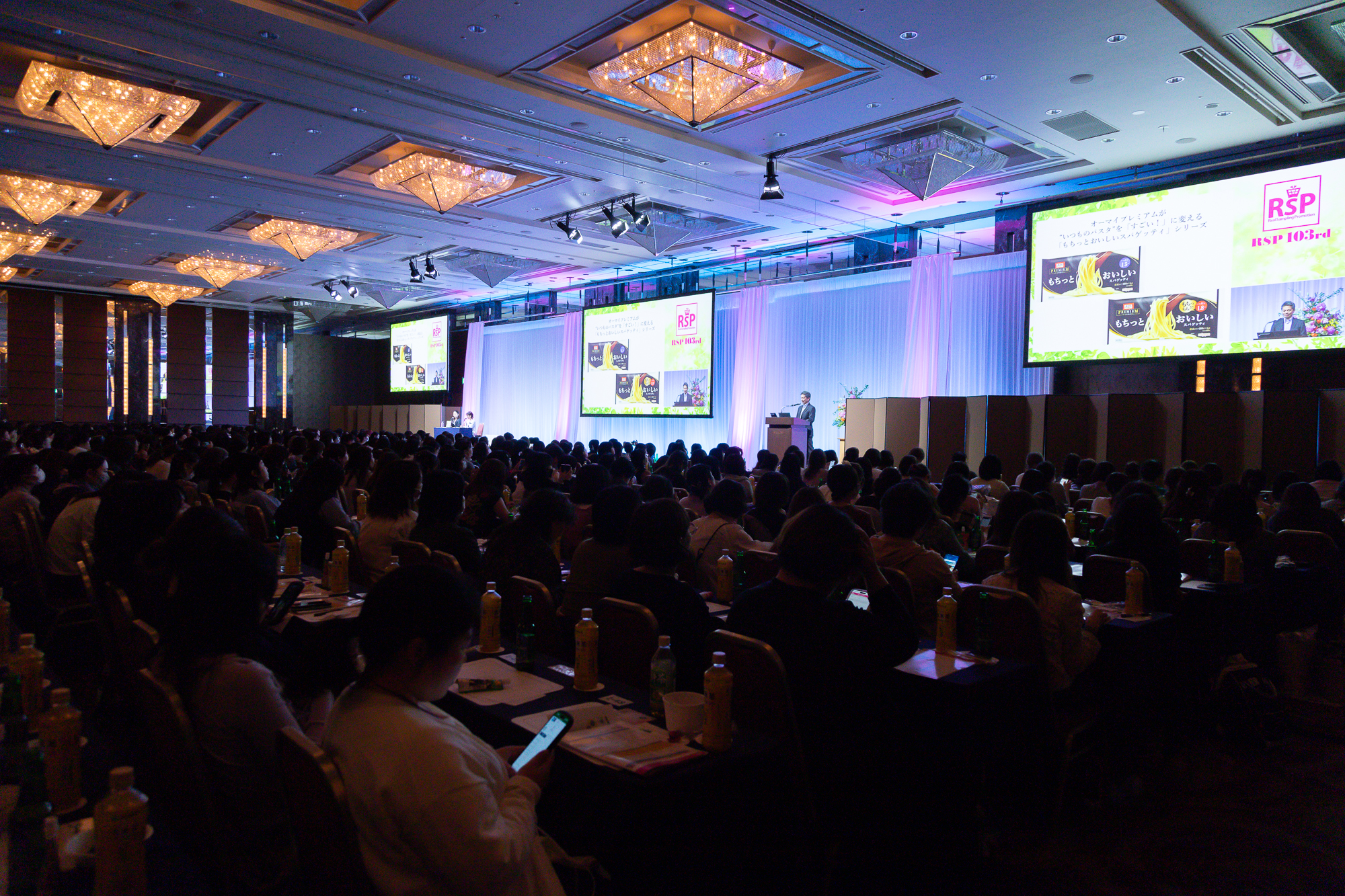 リアル開催された「リアルサンプリングプロモーション103rd（RSP103rd）」の会場の様子1枚目。暗めの会場で多くの参加者が同じ方向を向き椅子に着席し参加している。参加者が向く正面には大きいスクリーンが3つと登壇のための舞台があり、スクリーンには商品紹介の映像や登壇者が投影されている。