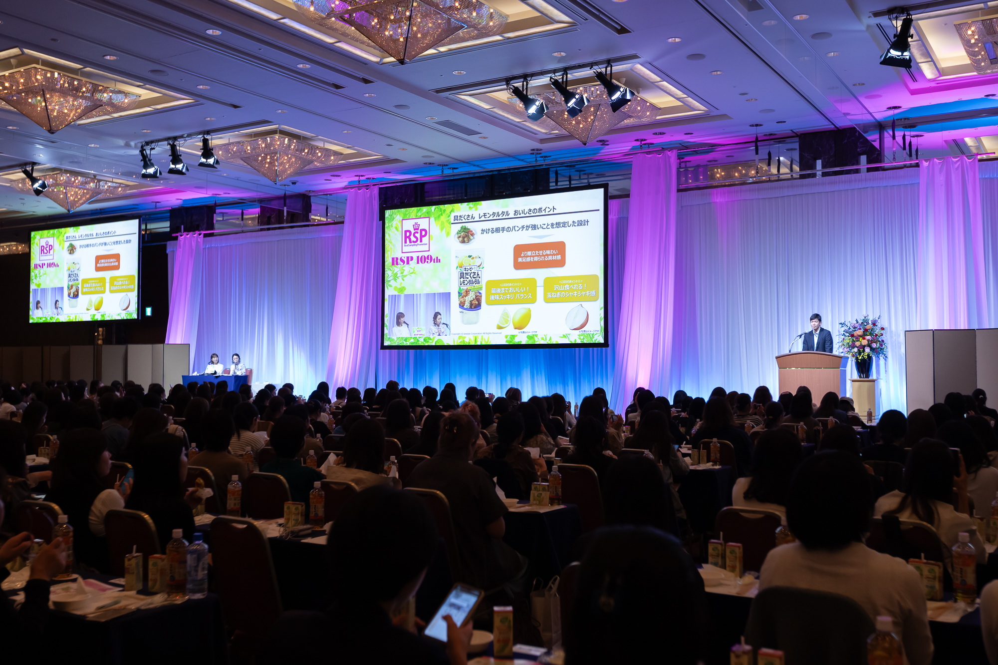「RSP（リアルサンプリングプロモーション）109th」のメイン会場の様子1枚目。暗めの会場で多くの参加者が同じ方向を向き椅子に着席し参加している。参加者の前には長机が用意されている。参加者が向く正面には登壇のための舞台があり、舞台には大きいスクリーンが2つと、登壇者が2名と司会者が1名。スクリーンには商品紹介の映像や登壇者が投影されている。