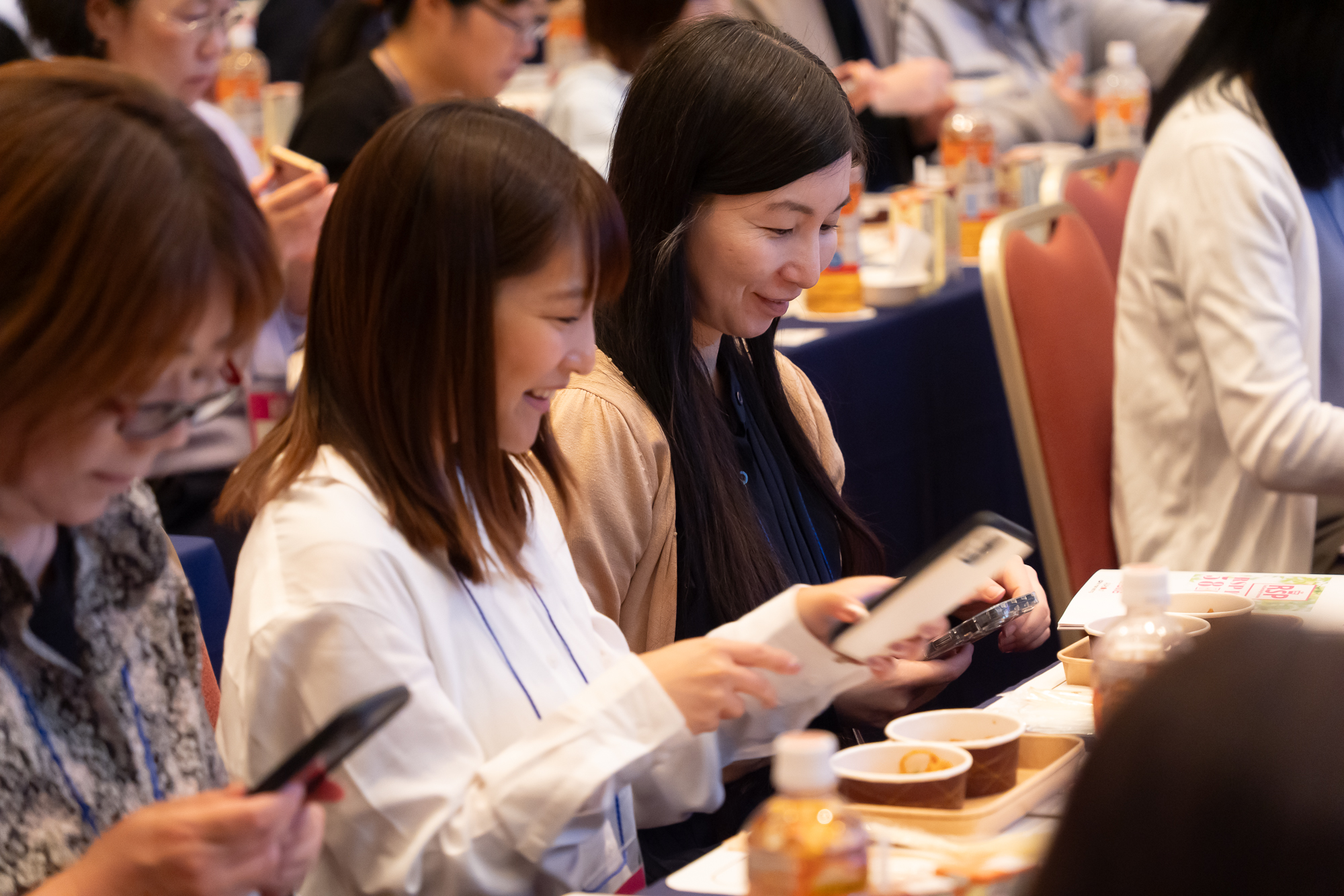 メイン会場の様子3枚目。参加者側の様子。着席した参加者2名がアップで写されていて、サンプル品を眺めたりスマホで写真を撮ったりしている。笑顔で楽しそうな様子。