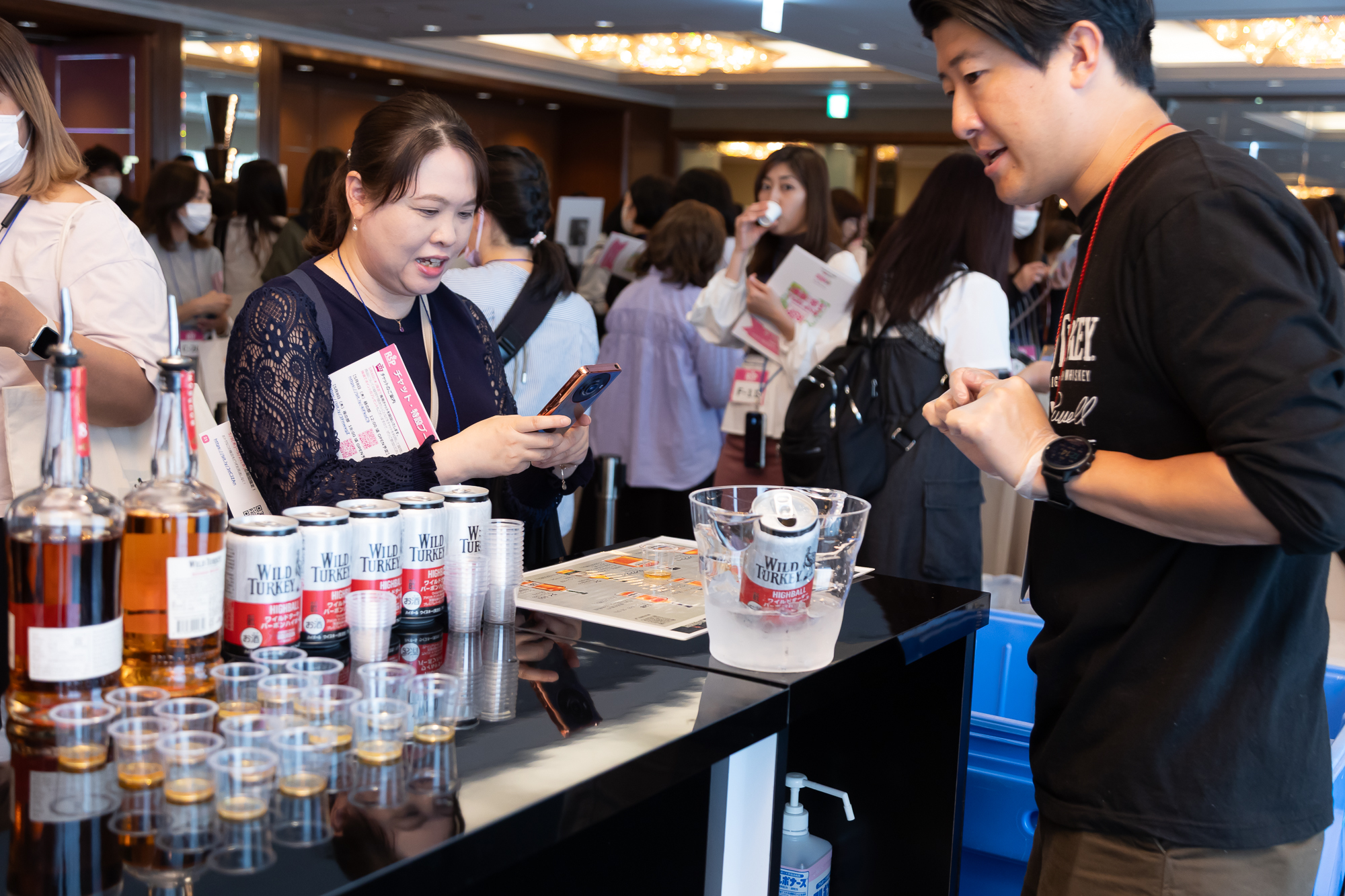 ブース会場の様子1枚目。お酒のサンプリングブースの様子。試飲用のコップやお酒の缶などが並び、ブース担当者が参加者に商品の説明をしている様子。