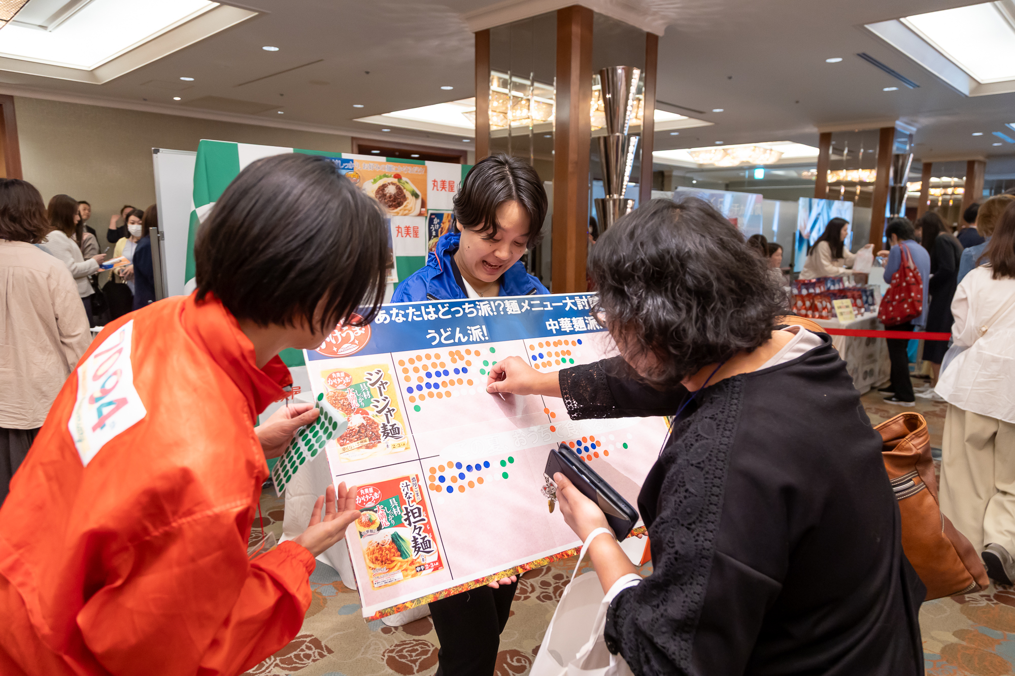 ブース会場の様子2枚目。「あなたはどっち派？」のシールを貼る方式の簡易アンケートを実施している。サンプル商品は「ジャージャー麺」と「汁無し担々麺」用のかけるソース。「うどん？それとも中華麺？どちらにかける？」というアンケートの様子。