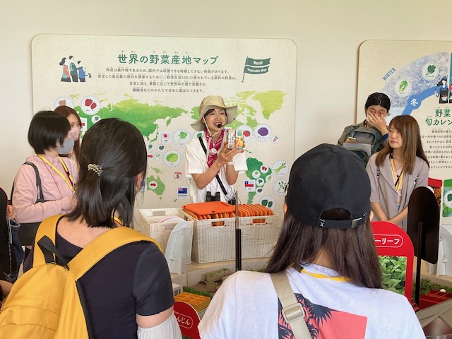 イベント当日の様子。「世界の野菜産地マップ」を背にして麦わら帽子を被ったガイドから野菜ジュース人について説明されている様子の参加者たち。